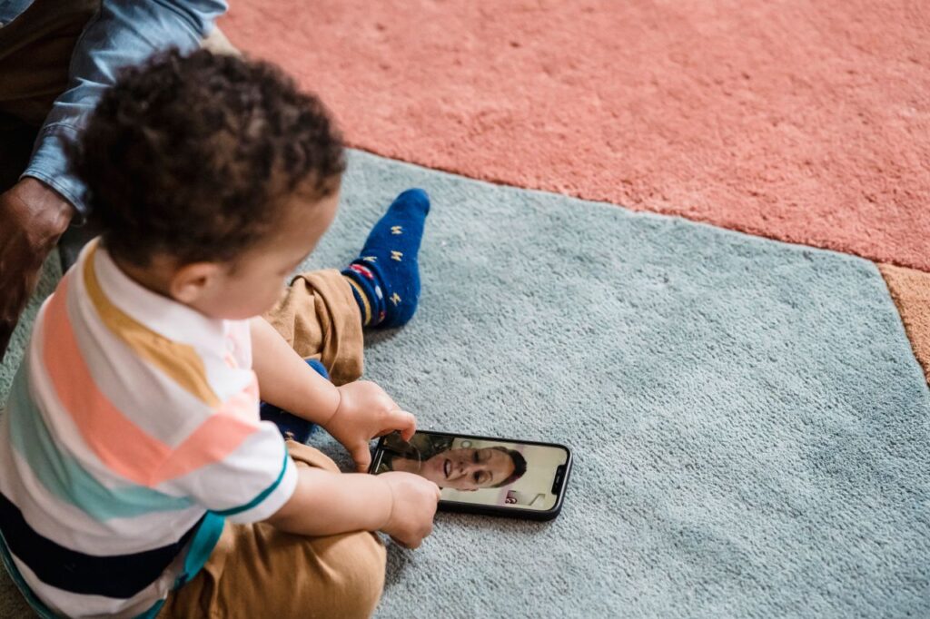 Child making a video call using a smartphone while sitting on a colorful carpet indoors.